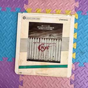 May include: A VHS video cassette cover for the horror film "C.U.J.O." based on the Stephen King novel. The cover features a white picket fence with red blood dripping down the fence posts. The text on the cover reads "Now there's a new name for terror."