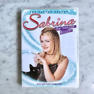 May include: DVD case for "Sabrina the Teenage Witch: The Second Season." The cover features a photo of the actress, a black cat, and the show's title in red script. The background is light blue with star accents. The text "Some girls lead a magical life" is at the bottom.