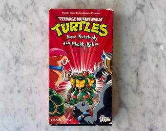 May include: A red and black VHS tape with a Teenage Mutant Ninja Turtles logo and the title "Super Rocksteady and Mighty Bebop". The image shows the four turtles in a fighting pose with two villains in the background.