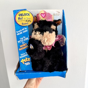 May include: A black and tan plush dog wearing a pink crown and a pink and purple sequined dress. The dog is holding a pink heart in its paw. The dog is packaged in a blue box with the text "UNLOCK Me! In Your NINTENDODS Game!" and "Petz DOGZ PACK".