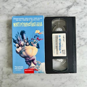 May include: A VHS tape and its case for the movie "Monty Python and the Holy Grail." The case is blue with a colorful illustration of the film's characters. The VHS tape is black.