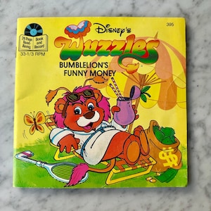 Könnte beinhalten: Ein gelbes Buch mit einem Comic-Löwen, der eine Sonnenbrille und ein weißes Hemd trägt, entspannt sich auf einem Liegestuhl mit einem Sack Geld. Das Buch trägt den Titel "Disney's Wizzles: Bumblelion's Funny Money" und hat ein Schallplattenspieler-Symbol auf dem Cover.