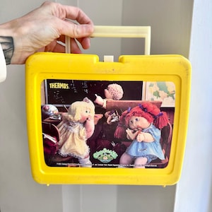 May include: Yellow vintage Cabbage Patch Kids lunchbox with a cream-colored handle. The lunchbox features a colorful image of two Cabbage Patch Kids dolls in a classroom setting. The word "THERMOS" is visible on the top left corner of the image.