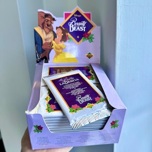 May include: A display box of Disney's Beauty and the Beast trading card packs. The box is lavender with rose and leaf accents. Each pack is white with purple text and the Beauty and the Beast logo. The box art features Belle and the Beast.