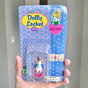 May include: A vintage Dolly Locket toy package. The packaging is purple with a repeating pattern of white and teal dots. The package features the words "Dolly Locket" in yellow and blue, and includes a small toy figure in a clear plastic case.