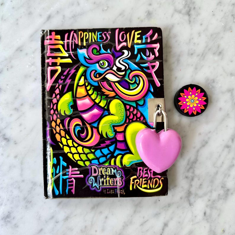Może przedstawiać: Czarny notatnik z kolorowym wzorem smoka, napisami "Happiness Love" i chińskimi znakami. Obecna jest r&oacute;wnież r&oacute;żowa kł&oacute;dka w kształcie serca i okrągła przypinka z kwiatem. Notatnik jest oznaczony jako "Dream Writers Best Friends".