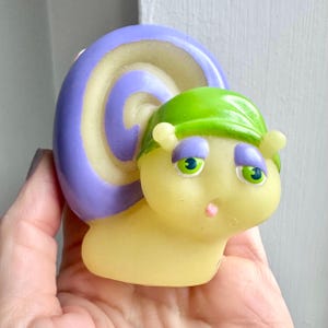 Puede incluir: Una caprichosa figura de caracol amarillo con una concha morada y crema y una gorra verde. El caracol tiene ojos verdes, párpados morados y boca rosa. La figura se sostiene en una mano.