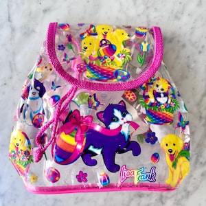 May include: Clear plastic backpack with a pink trim and drawstring closure. The backpack is decorated with colorful illustrations of puppies, kittens, butterflies, and Easter eggs. The text "Lisa Frank" is visible on the lower right.