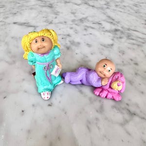 May include: Two small figurines featuring a blonde doll in a teal dress with a purple sash and a bald baby doll in a purple onesie lying next to a pink dog. The blonde doll holds a small white book with the text "LITTLE BOOK" printed on the cover.