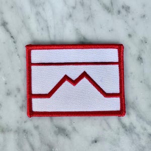 May include: A rectangular embroidered patch featuring a white background with a bold red border. The design includes a horizontal red line across the top and a central red mountain-like peak shape that connects to the bottom border.