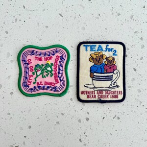 Puede incluir: Dos parches bordados. Uno es rosa con ribete verde, con ranas y el texto "Let's Go To The Hop B.C. Dance Father-Daughter". El otro es color crema con borde azul, que representa osos en una taza de t&eacute; y el texto "Tea for 2 Mothers and Daughters Bear Creek 1989".