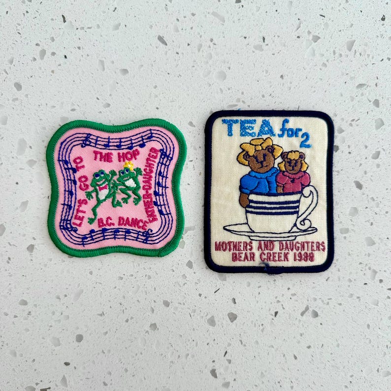 Puede incluir: Dos parches bordados. Uno es rosa con ribete verde, con ranas y el texto "Let's Go To The Hop B.C. Dance Father-Daughter". El otro es color crema con borde azul, que representa osos en una taza de t&eacute; y el texto "Tea for 2 Mothers and Daughters Bear Creek 1989".