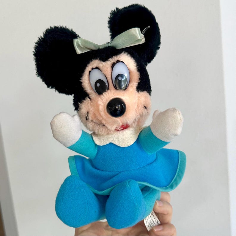 May include: A plush toy of Mickey Mouse's girlfriend Minnie Mouse. She is wearing a blue dress with a white collar and a green bow. She has black ears and a pink nose.