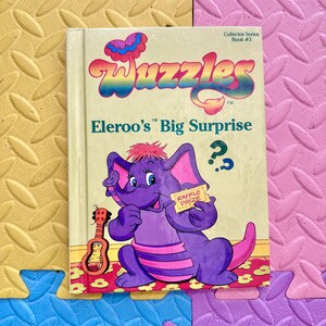 Op de afbeelding: Een vintage kinderboek getiteld "Wuzzles: Eleroo's Big Surprise". De cover toont een paars olifantenpersonage met een loterijprijs, een kleine gitaar en de tekst "Collector Series Book #3."