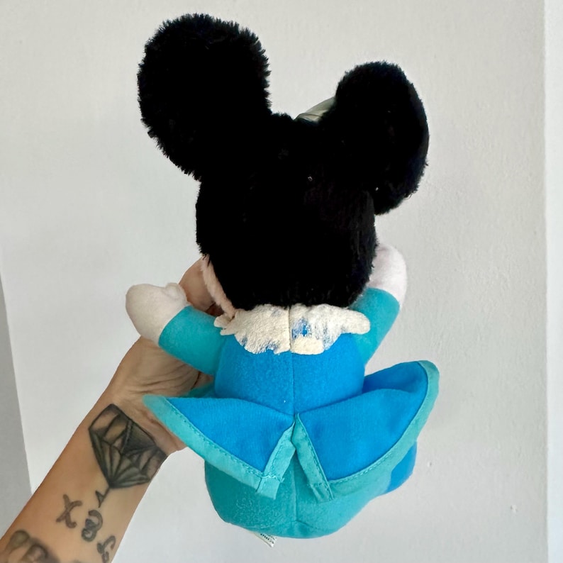 May include: A plush toy of Mickey Mouse wearing a blue and white outfit with a white collar. The toy is sitting on a person's arm.