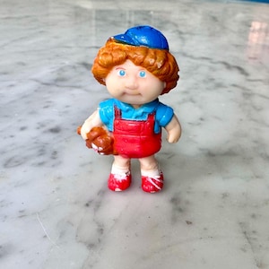 Könnte beinhalten: Vintage Puppenfigur mit orangefarbenem Haar, blauen Augen und einer blauen Kappe. Die Puppe trägt ein blaues Hemd unter einer roten Latzhose und roten Schuhen. Sie hält einen kleinen braunen Teddybären.