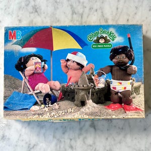 May include: A 100 piece Cabbage Patch Kids jigsaw puzzle featuring three Cabbage Patch Kids dolls on a beach. One doll is wearing a swimsuit and sitting in a beach chair, another doll is wearing a swimsuit and building a sandcastle, and the third doll is wearing a swimsuit and flippers and holding a snorkel.
