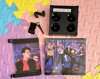 May include: A black plastic toy speaker with the words "NEW KIDS" on the front. The speaker has four black speakers on the front. There is a black and white photo of a boy band on a white background. The photo shows five boys in different poses. The photo is framed in black plastic.