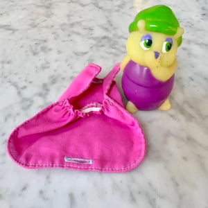 May include: A small, yellow, rubber-like creature figurine with a green hat, purple body, and green eyes, positioned next to a bright pink fabric garment featuring a gathered neckline, thin shoulder straps, and a single stitched buttonhole.