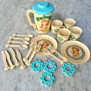May include: A vintage Cabbage Patch Kids themed children's tea set. The set includes a pitcher, cups, plates, utensils, and flower-shaped cookie cutters. The pitcher and plates feature the Cabbage Patch Kids logo and illustrations.