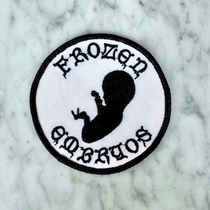 Op de afbeelding: Een ronde geborduurde patch met een witte achtergrond en een zwarte rand. Het midden toont een zwart silhouet van een foetus. De tekst "FROZEN" staat in een boog bovenaan en "EMBRYOS" onderaan in een gotisch lettertype.