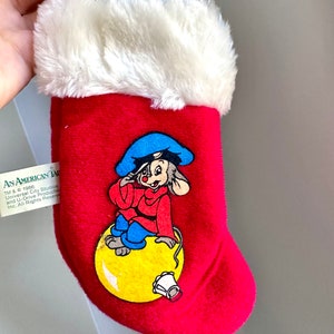 Peut inclure: Chaussette de Noël en peluche rouge avec un revers en fausse fourrure blanche. La chaussette présente une illustration de dessin animé d'une souris portant un chapeau bleu et une veste rouge, assise sur une boule jaune avec une cloche.