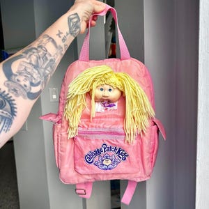 May include: A pink nylon backpack featuring a Cabbage Patch Kids doll face with yellow yarn hair attached to the front. The front pocket displays the Cabbage Patch Kids logo in blue and purple text above a graphic of a cabbage leaf.