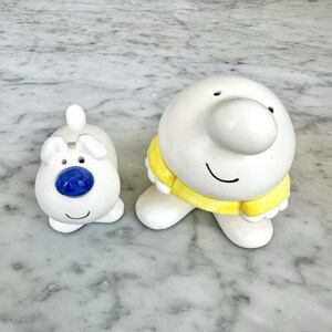 May include: A ceramic salt and pepper shaker set featuring a small white dog with a blue nose and a larger round white character wearing a yellow shirt. Both figures have simple black line smiles and smooth, glossy white surfaces.