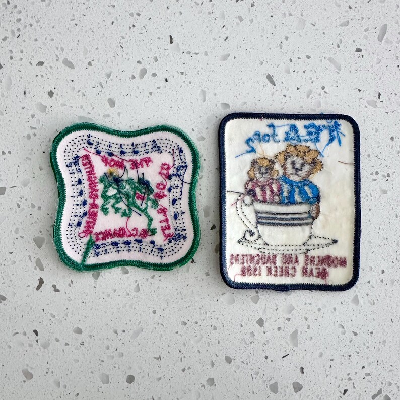 Puede incluir: Dos parches bordados. Uno es cuadrado con borde verde y centro rosa, con texto y un dise&ntilde;o verde. El otro es rectangular con borde azul marino, que representa dos figuras en una taza de t&eacute; con texto arriba y abajo.