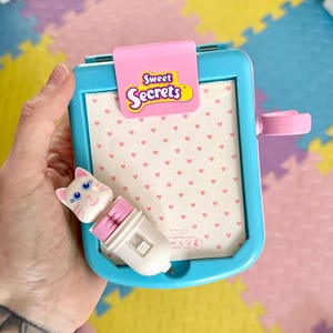 May include: A blue and pink plastic toy notepad with a white cat-shaped pen. The notepad has a pink flap with the words "Sweet Secrets" on it. The notepad has a white background with a pink heart pattern.