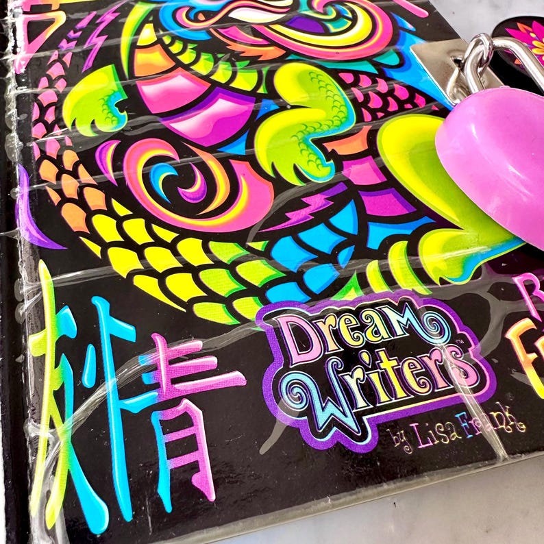 Może przedstawiać: Czarny notatnik Lisa Frank "Dream Writers" z kolorowym wzorem smoka. Smok ma żywe r&oacute;żowe, ż&oacute;łte, niebieskie i zielone akcenty. Notatnik ma spiralną oprawę i r&oacute;żową zawieszkę w kształcie serca.