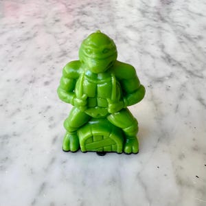 May include: A green plastic figurine of a Teenage Mutant Ninja Turtle character. The figure is in a fighting stance, holding two weapons. The figure is sitting on a turtle shell base. The figurine is on a white marble surface.