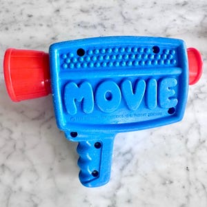May include: A vintage blue toy video camera with the word "MOVIE" in white letters. The camera has a red lens and a handle. The text "© 1976 MATTEL, INC. MEXICO, U.S. PATENT PENDING" is printed on the camera.