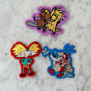May include: Three embroidered patches featuring cartoon characters. The top patch shows a brown beaver and a yellow creature with a pink border. The bottom left patch depicts a boy with yellow hair in a red border. The bottom right patch shows two characters in a blue border.