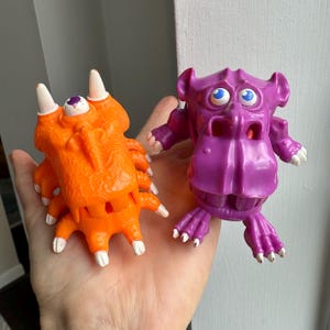 Peut inclure: Deux jouets monstres vintage colorés. L'un est orange avec plusieurs pattes et des griffes à bout blanc. L'autre est violet, ressemblant à un hippopotame avec des yeux bleus et des griffes à bout blanc. Les deux sont tenus dans une main.