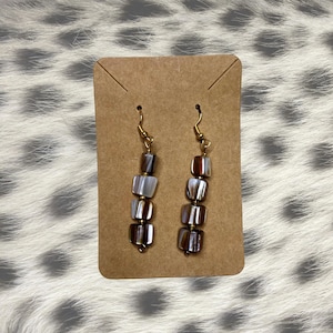 以下が含まれることがあります： ゴールドカラーのフックと、茶色、グレー、白の色合いの正方形のイリデッセントビーズが縦に並んだドロップイヤリングのペア。イヤリングは茶色のカードに表示されています。