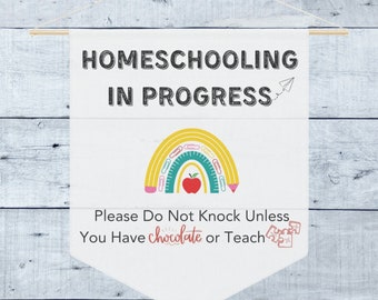 May include: A white banner with the text "HOMESCHOOLING IN PROGRESS" and a rainbow made of pencils, a ruler, and paper clips. The banner also says "Please Do Not Knock Unless You Have Chocolate or Teach".