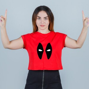 Puede incluir: Una mujer con un top rojo corto con un dise&ntilde;o de m&aacute;scara negra de Deadpool. Est&aacute; apuntando con los dos dedos &iacute;ndices hacia arriba.