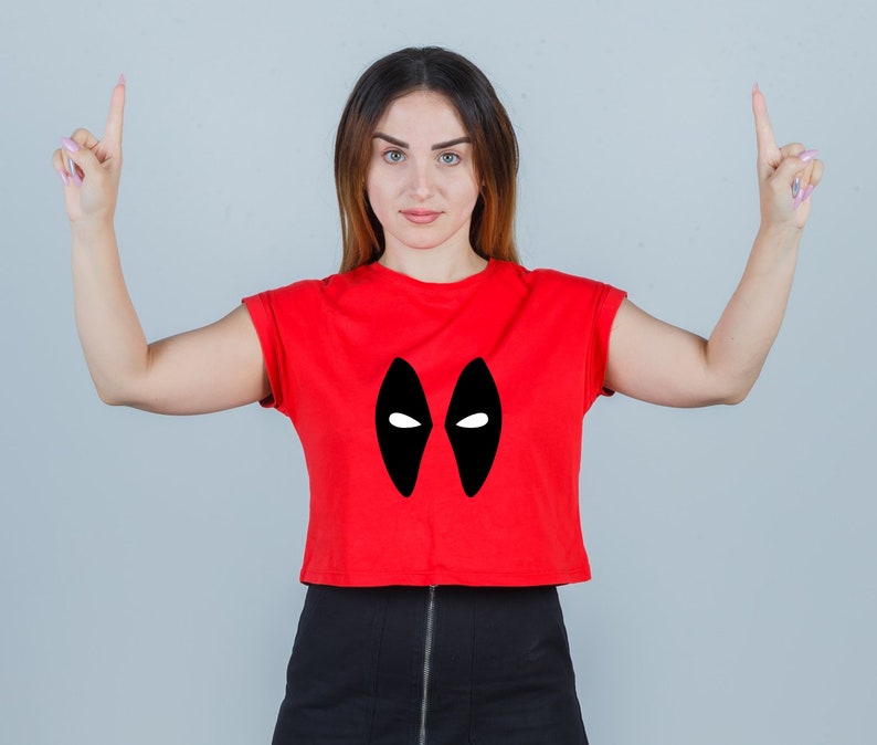 Puede incluir: Una mujer con un top rojo corto con un dise&ntilde;o de m&aacute;scara negra de Deadpool. Est&aacute; apuntando con los dos dedos &iacute;ndices hacia arriba.