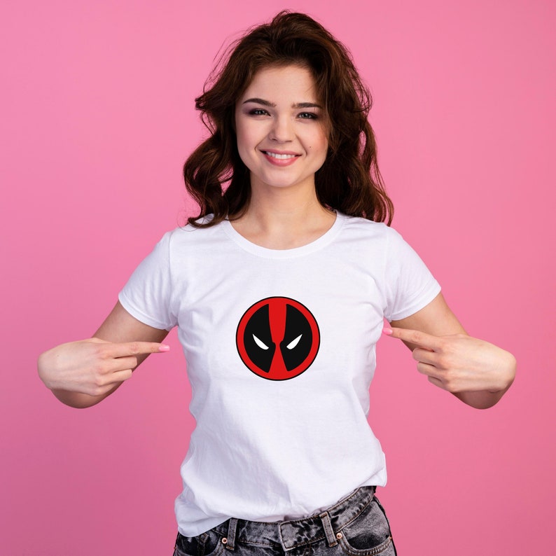 Puede incluir: Camiseta blanca con un logotipo de Deadpool rojo y negro en la parte delantera.