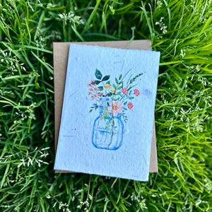 May include: A handmade card with a watercolor illustration of a blue glass jar filled with a bouquet of pink, white, and yellow flowers. The card is on a green grass background.