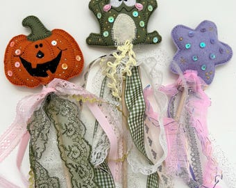 May include: Three felt wands featuring a pumpkin, a frog, and a star. The pumpkin is orange with a black face, the frog is olive green with white eyes, and the star is lavender. Each wand has a wooden handle adorned with various ribbons, lace, and sequins.
