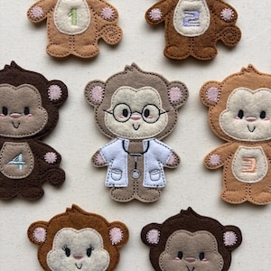 May include: Felt monkey appliques in various shades of brown, tan, and cream. Some monkeys have numbers embroidered on their bellies, while one is dressed as a doctor with glasses and a stethoscope. The appliques are designed with pink accents.