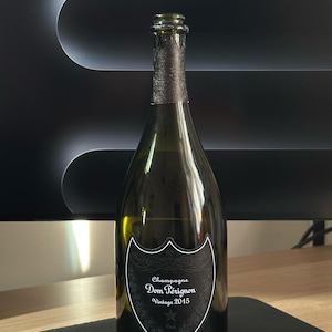 Op de afbeelding: Een groene Dom Pérignon Champagne fles, Vintage 2015, met een zwart-wit etiket. De fles heeft een donkere folie rond de hals en wordt gepresenteerd op een donkere ondergrond.