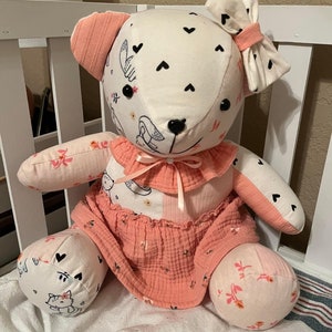 May include: A handmade plush bear with a cream-colored head and limbs, adorned with black hearts and floral patterns. The bear wears a peach-colored dress and bow, and has black button eyes. The bear is sitting on a white surface.