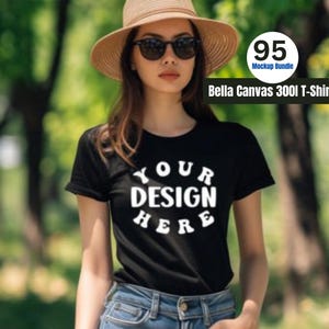 May include: A black t-shirt with white text that says "YOUR DESIGN HERE" on the front. The t-shirt is being worn by a person with brown hair and sunglasses. The person is wearing a brown hat and blue jeans.