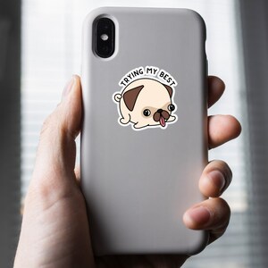 May include: A sticker featuring a cartoon pug with a tan body, brown ears, and a black nose. The pug has its tongue sticking out and the text "TRYING MY BEST" in a circle above it. The sticker is on a gray phone case.