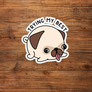 May include: A sticker featuring a cartoon pug with a light tan body, brown ears, and a pink tongue sticking out. The text "TRYING MY BEST" is arched above the pug's head in black letters. The sticker is on a wooden background.