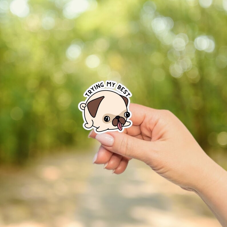 May include: A sticker featuring a cartoon pug with a tan body, brown ears, and a black snout. The pug has its tongue sticking out and the text "TRYING MY BEST" above it. The sticker is held in a hand against a blurred green background.