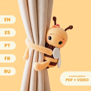 Pode incluir: Um prendedor de cortina de abelha de crochê em tons de amarelo, marrom e branco, abraçando uma cortina bege. A abelha tem olhos grandes e uma expressão sorridente. O texto "crochet pattern PDF + VIDEO" é visível.